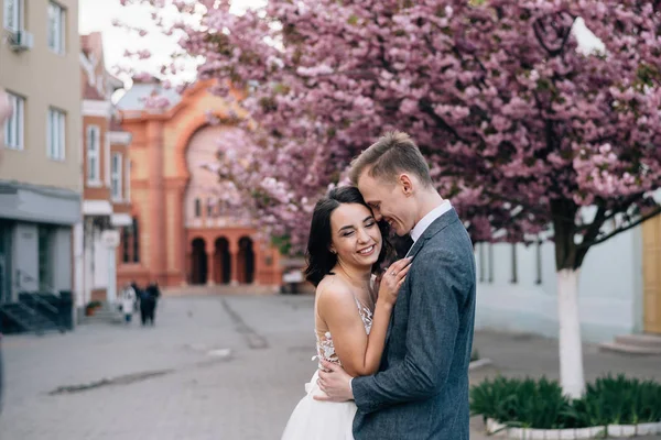 Nevěsta a ženich se smějí na ulici. Svatební šaty a ženichův oblek — Stock fotografie