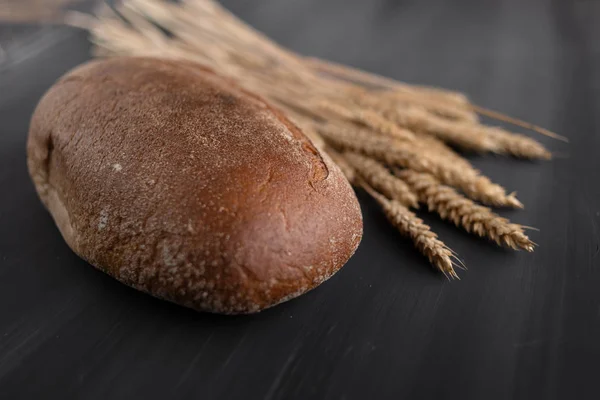 Una hogaza de pan oscuro sobre la mesa, junto a ella está el trigo en una espiga — Foto de Stock