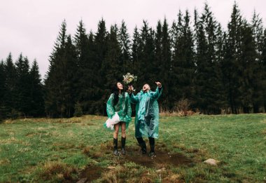 Gelin ve damat yağmurluk giyiyorlar, su birikintisine atlıyorlar ve neşeyle gülüyorlar.