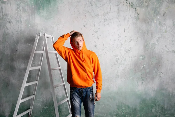 Молодой человек стоит рядом с лестницей снимает капюшон на фоне старой стены . — стоковое фото