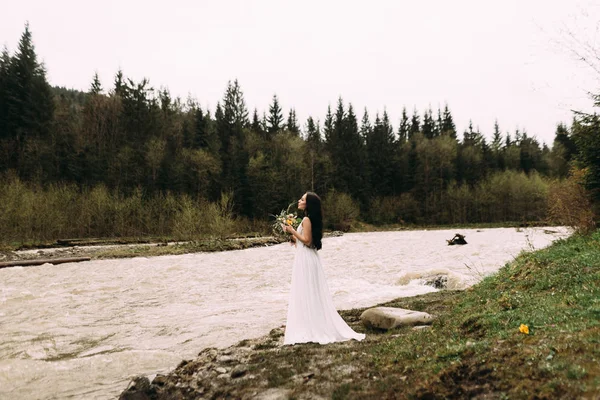 Novia el día de la boda, está en la orilla del río. Hermoso ramo de boda en sus manos — Foto de Stock