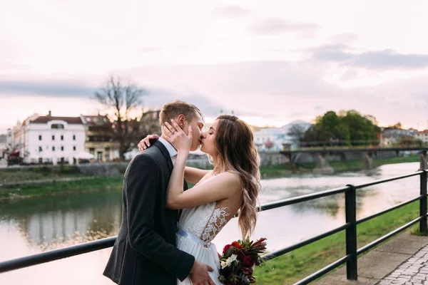 Bruden omfamnar brudgummen och kysser honom på stranden av floden. — Stockfoto