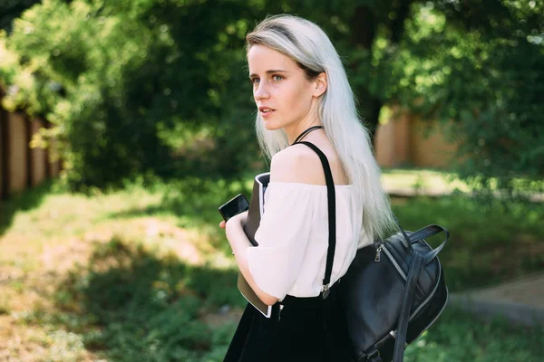Chica se para de lado sosteniendo un gadget y una revista en sus manos y una mochila negra . —  Fotos de Stock