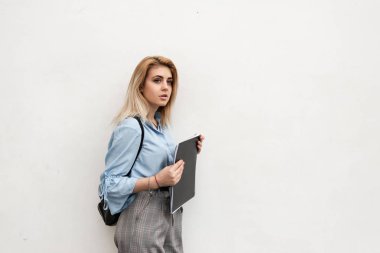 beyaz bir duvarın arka planında siyah bir defter ve plache üzerinde bir rukzak tutan bir kız duruyor.