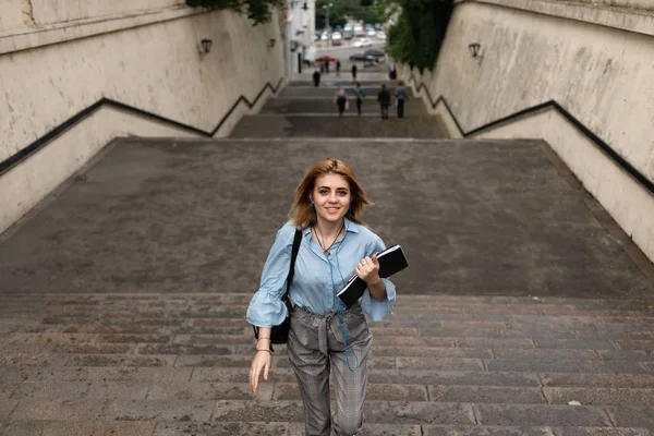 Estudiante satisfecho camina arriba sosteniendo un cuaderno y una mochila mientras escucha música agradable . — Foto de Stock