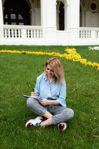 Una estudiante feliz se sienta en un césped en un parque. Escribe en un cuaderno un ensayo . — Foto de Stock