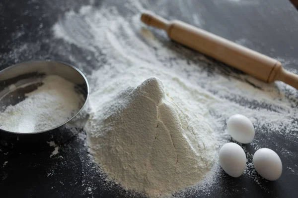 Still life handful of flour on a black background, rolling pin, chicken eggs and a sieve are nearby — Stock Photo, Image