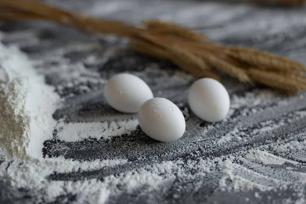 On a black background a pile of flour, chicken eggs, an ear of wheat — Stock Photo, Image