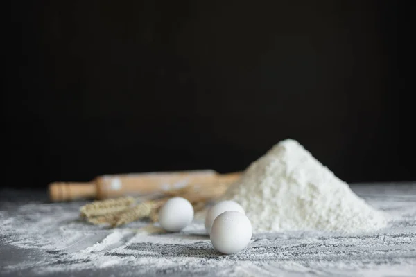 Three chicken eggs on a black background, a handful of white wheat flour rolling pin. — Stock Photo, Image