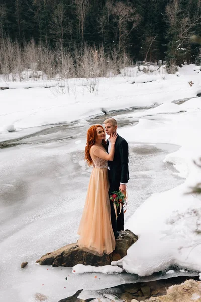Boda en invierno, la novia y el novio están de pie en la orilla de un río congelado y abrazándose. Elegante pareja enamorada — Foto de Stock