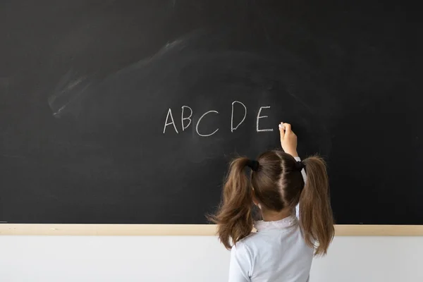 授業中に学校で甘い女の子 黒い板の前に子供が立っている 彼女は英語でチョークでアルファベットの文字を書きます 碑文の場所です 教育の概念 — ストック写真