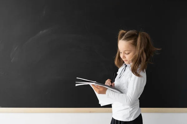 学校での美人女子生徒 彼女はきれいな黒い板の近くに立って 学校のノートに書いています 彼女は制服を着ている 碑文の場所です 教育理念 — ストック写真