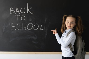 Mutlu liseli kız tahtaya yakın duruyor. Omuzlarında bir sırt çantası vardı. Okuldayken başparmağınla işaret et. Boş alan