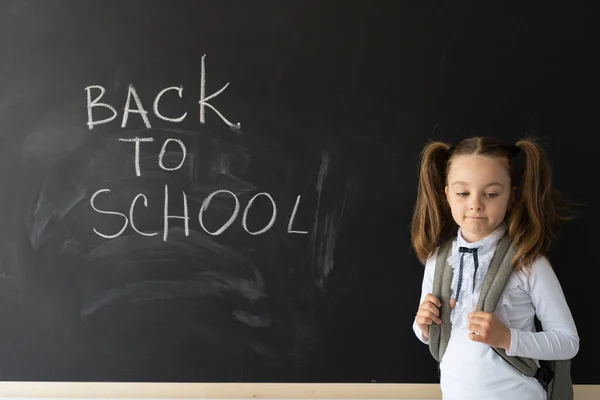 sad schoolgirl stands near the blackboard. The backpack is put on shoulders. the boring beginning of the school year. Place for text