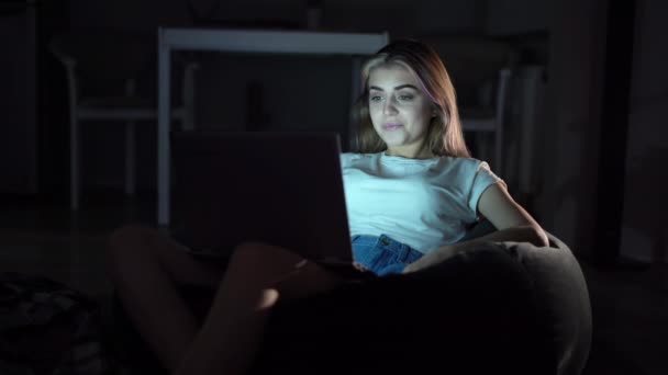 Bella ragazza seduta su una sedia e guardando un film su un computer portatile. E 'sola in una stanza buia. Cinema online. Guardare video a casa . — Video Stock