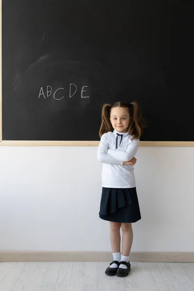 Porträtt av en skolflicka med flätor, hon står nära svarta tavlan. Engelska bokstäver skrivs av ett barn. Ort för text. — Stockfoto