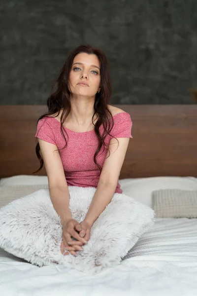 A lovely young brunette girl woke up in the morning in her bedroom. Her hair is loose and her face is sad. She is sitting on the bed and laid her hands on a large white pillow. Pink top T-shirt fits