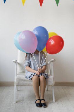 Doğum gününde kucağında çok renkli balonlarla dolu bir odada beyaz bir koltukta oturan tatlı küçük kız. Kendini sakladı. Yüzü görünmüyor. Tatilin atmosferi. Genç bir bayan için mutluluk. Ev kutlama konsepti. Beyaz arka plan