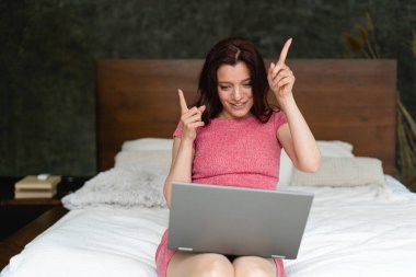 Pijamalı genç bir kadın odada yatakta oturmuş el kol hareketi yapıyor. Ayaklarının üzerinde bir dizüstü bilgisayar var. Evdeki web kamerası iletişimi. Genç bir bayanın hayatıyla ilgili video çağrısı. İletişimdeki bilgisayar teknolojisi kavramı.