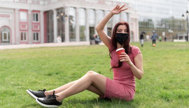 Hastalıklara, alerjilere veya hava kirliliğine karşı korumak için siyah maske ve tıbbi maske takan genç bir kadın parkta çimlerin üzerinde oturuyor. Arkadaşlarına el sallıyor. Salgın sırasında mesafeyi koruma kavramı