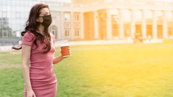 公園の芝生の上には 黒いマスクと病気 アレルゲン 大気汚染から守るための医療用マスクを着た若い女性が立っています 自然の中でコーヒーを飲む テキストの場所 — ストック写真