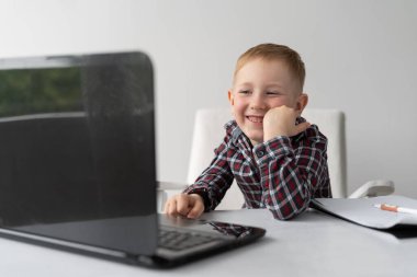Bir okul çocuğu evde dizüstü bilgisayarın başında oturuyor ve gülümsüyor. Online dersler. Karantinada çocukların eğitimi. Web kamerasında ödev yapıyorum. Mesafe öğrenme ve yaşam tarzı. Bir okul çocuğunun kendini tecrit etmesi. Metin için bir yer.