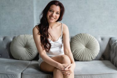 Oturma odasındaki kanepede siyah saçlı ince bir kız model oturuyor. Saten pijamalarıyla poz veriyor. Kız gülümsüyor ve flört ediyor. Mankenlik işi. Ev sateni kostümü. Dairelerdeki güzel esmerleri düşün..