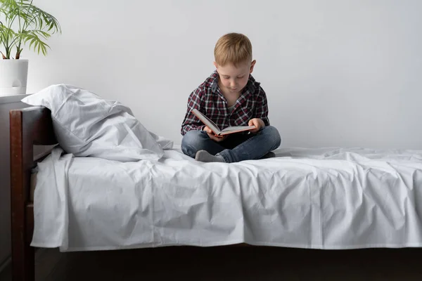 A boy on self-isolation reads a book and stays at home. He is sitting on the bed and his legs are folded. Self study lessons. Rest student from school. Free time and education. Place for text.