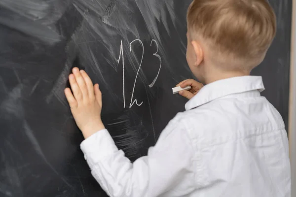 A boy stands with his back in front of a blackboard at school. Math lesson in elementary school. A schoolboy at the blackboard writes numbers in chalk. Child\'s hand lies on a blackboard. Education of primary schoolchildren. Place for text.