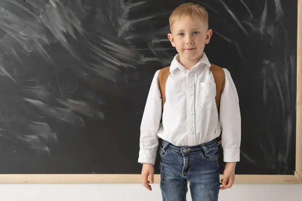 The younger schoolboy is standing near the blackboard. Portrait of a joyful boy in a shirt and jeans with a school bag. School. A clean blackboard. Place for text. Back to school.