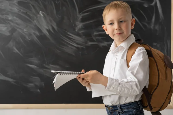 学校の黒板の近くの少年の肖像画 その少年は大きなノートを手に持っている 彼は驚いている 後ろに茶色のスクールバッグがあります 小学校での教育の概念 学校に戻る テキストの場所 — ストック写真