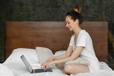 Otel odasında çalışan, beyaz çarşaflı bir yatakta oturan ve dizüstü bilgisayarla çalışan modern bir genç kadın. Kızın saçı kafasında toplanır ve kıvrılır. Hafta sonu sabahı ve kişisel bilgisayarın başında bir iş kadını. Uzaktan kumandalı iş ve bir kadın. Uzak