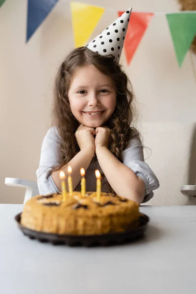 6-7 yaş arası güzel bir kızın doğum günü. Saçları kıvırcık ve kafasında şenlik şapkası var. Aydınlık bir odada, çok renkli bayraklar asılı ve bir masa standı var. Mumlu bir doğum günü pastası var. Bebek yumruklarıyla başını destekliyor. 