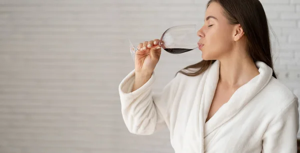 A young Caucasian woman is relaxing in a hotel room. Hold a glass of red wine in hands. Tasting a drink. Place for text. — Stock Photo, Image