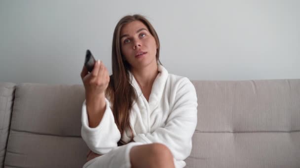 Mujer joven viendo televisión en casa, sentada en el sofá. Somnoliento y cansado, bostezando. Cambia un canal — Vídeos de Stock