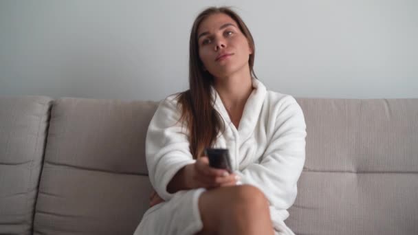Een jonge vrouw die thuis op de bank tv zit te kijken. Selecteer een film om 's avonds naar te kijken. Witte badjas. — Stockvideo