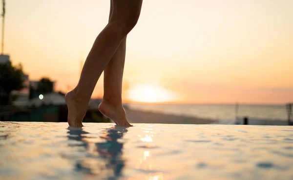 Female feet in the water on a sunset background. Slender legs barely touch the water. In the foreground, a step along the water is visible. Behind the sun is visible on the horizon. Glimpses of the sun on the water.