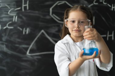 Sınıftaki kimya dersinde tahtaya yakın şirin bir kız. Elinde mavi sıvı olan şişeyi tut. Güvenlik gözlükleri ve beyaz önlük.