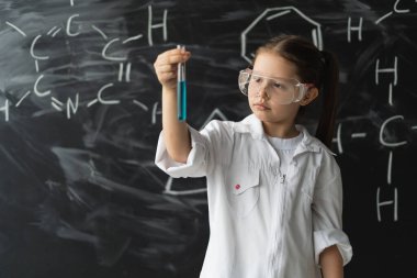 Gözlüklü ve beyaz önlüklü küçük kız öğrenci tahtaya yakın duruyor. Test tüpünü sıvıyla kaldırır ve bakar. Kimya dersi konsepti
