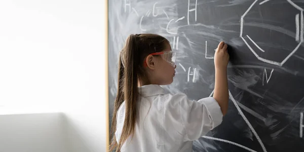 クラスの化学レッスンで黒板に数式を書くかわいい女の子 白いコートとゴーグル コピースペース — ストック写真