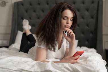 Yatakta yatarken cep telefonuyla mesajlaşan güzel bir genç kadının görüntüsünü kapat. Genç esmer, modern yatak odasında cep telefonunu kullanıyor..