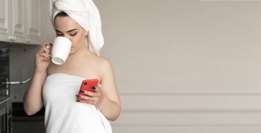 Çok güzel bir genç bayan mutfakta durup kahve içerken cep telefonu kullanıyor. İnternet sörfü, bağımlılık, aygıt konsepti, metin için alan, kopyalama alanı