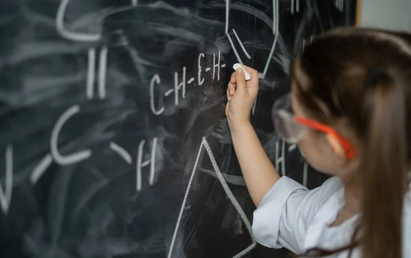 黒板の近くの女子高生は化学の問題を解決する 要素の数式を書く 白いコートとゴーグル オフライン教育の概念 — ストック写真