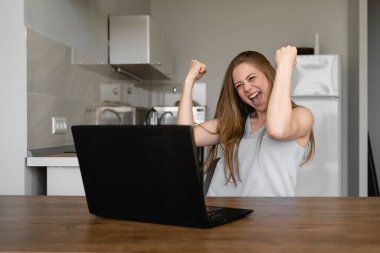 Bilgisayarıyla uzaktan çalışan heyecanlı güzel bir kadın. Mutfak arka planında alkışlayarak ve yumruklarını havaya kaldırarak başarısına seviniyor.