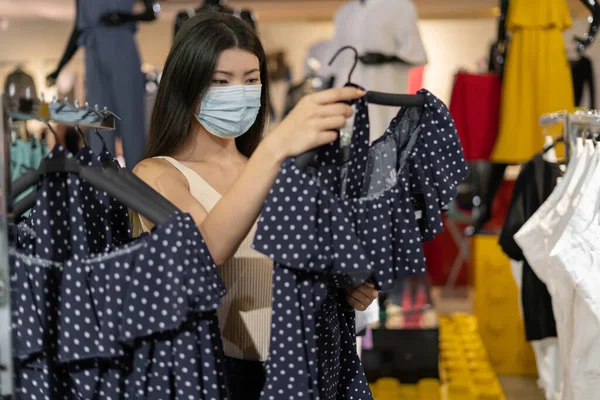 young beautiful korean woman dressed in a protective medical mask on her face stands in a clothing store. Buying things during a pandemic.