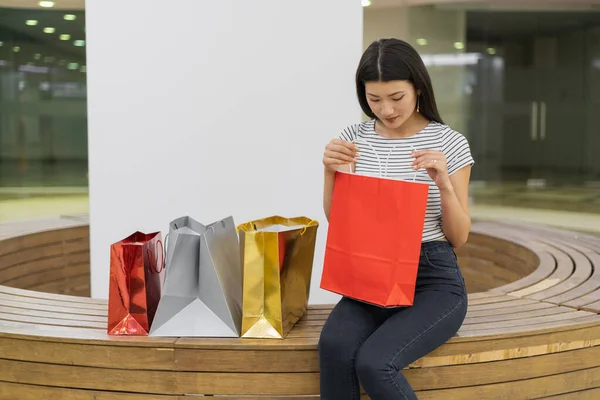 一位年轻妇女坐在购物中心的长椅上 拿着一个红色纸袋 看里面 — 图库照片