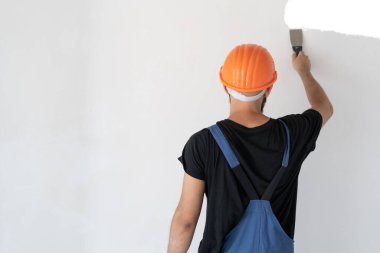 Erkek müteahhit özel kıyafetler giyer ve turuncu bir şapka beyaz bir duvarın yanında durur. Duvara macun dök. Spatula elinde. Çalışma süreci. Boşluğu kopyala