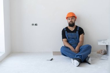 Müteahhitte dinlenin. Mavi üniformalı ve turuncu şapkalı sakallı bir adam mola sırasında yerde oturuyor. Mesaj için yer. Boşluğu kopyala.