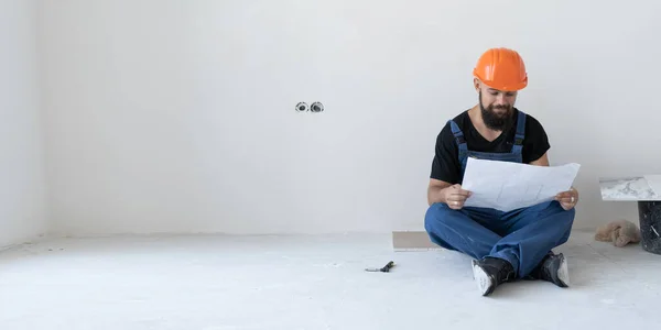 A brutal male builder is sitting on the floor. Dressed in a blue uniform and an orange mask. He holds a shabby and old sheet with drawings in his hands. Place for text.