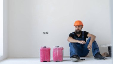 Müteahhitte dinlenin. Mavi üniformalı ve turuncu şapkalı sakallı bir adam mola sırasında yerde oturuyor. Boş alana bakıyor. Mesaj için yer. Boşluğu kopyala.